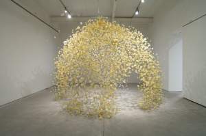 Return is a dome-shaped installation made of suspended yellow and white flower-like objects hangs in the center of a white-walled gallery space.