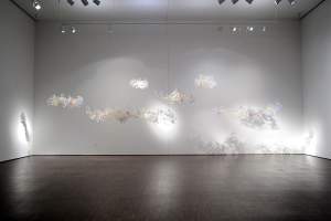 Cloud is an abstract installation composed of clusters of delicate, translucent cloud-like sculptures suspended in front of a white wall, illuminated by overhead spotlights, casting intricate shadows on the surface.