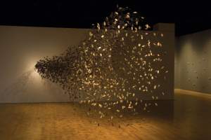 Web #5 is a large sculptural installation of many small, pod-like objects suspended in midair, fanning outward from a central point on a wall in a dimly lit gallery space, like a horizontal tornado.