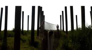 Eighteen posts are driven into a grassy incline in rows in this digital print titled We arrive at the port and we…. A person at the center of the photograph is backlit against the washed out sky. They bend or bow forward and hold a net up high with both hands.