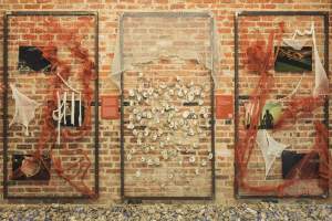 The bas du fleuve installation includes three vertically oriented metal frames hanging against a brick wall. Each frame is strung with fiber and ropes, nets, photographs, and oyster shells are suspended from the grids. Informational labels, illegible here, are between the panels, and the ground is piled with blue- and white-toned oyster shells.