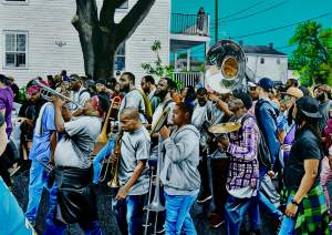 A few dozen people walk to our left in a dense procession in this painting titled WE OUTSIDE. One person plays a trumpet and another a tuba. Four others carry trombones and one a hi-hat cymbal. People are dressed casually and they look around. A house with white siding on the far side of the street and a high-keyed, turquoise-blue sky creates the background.