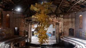 In a dilapidated theater with graffitied brick walls and exposed metal rafters, tendrils and swathes of golden paper cutouts cascade from the ceiling in tangles, forming a soft tornado form.
