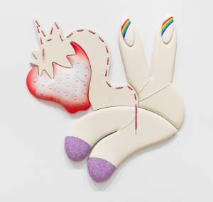 This shaped leather piece is titled Play Day. Two fingers lifted in a V to the right have long nails painted with rainbows. Below, two more finger-like forms curve to the left and down. Purple caps or socks patterned with blue are on the rounded tips. To the left, between the two pairs, the piece is the silhouette of a cartoon unicorn with a strawberry for a face.