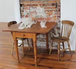This work, titled Archives, is made up of a wooden table and two wood chairs in front of a wall with partially exposed brick. At least five white scrolls on the table at first appear to be rolls of gauze.