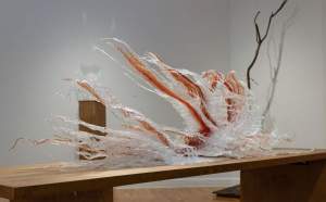This glass sculpture, titled Inherit, lies along a wooden table. Clear and frosty-white filaments create long, banner-like pages that lift up and flutter to our left in this view. Each banner is streaked with deep red along its center.
