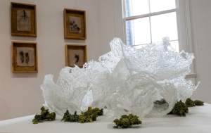 This glass sculpture, Pessaries, resembles slices of frosty foam that surge up and off a table top. Bunches of green leaves tied into bundles are scattered around the perimeter of the sculpture.
