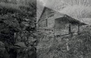 This black and white photo print, In Conversation from the SOIL series, juxtaposes a close up view of rocky dirt, rocks, and grass against a single-story wood house with a deep overhang over two empty doorways.