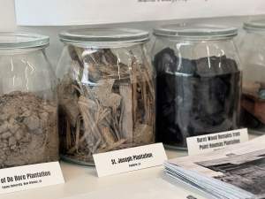 Four glass containers with glass lids are lined up against a white wall in this piece titled SOIL Jars. Labels in front of the two middle jars read, “St. Joseph Plantation, Vacherie, LA” and “Burnt Wood Remains from Point Houmas Plantation,” though the location on this second label is obscured by a stack of booklets. The visible part of the label on the leftmost jar reads “of De Bore Plantation, Tulane University, New Orleans, LA.”