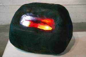 “Joey” is a sculpture of a lit automatic tail light set into a rounded biomorphic black form like a small boulder. The form has a spackled plaster-like texture and a matte finish.