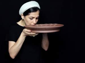 This still from a performance of “Bite” shows the artist Raheleh Filsoofi holding a large shallow unfired clay platter to her mouth. The edge of the platter shows a series of methodical, patterned bite marks as she works her way around the edge. She wears a white headwrap and a black shirt in a black space.