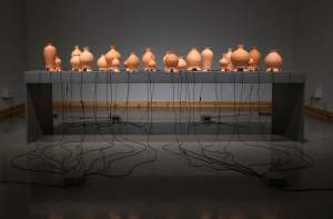 “The Inh(a/i)bited Space” is a sculptural installation of 64 bulbous clay vases of various scales staggered along the length of a long gray table. From openings of the base of each, black wires extend and drape down to the floor, where they connect at 4 electrical junctures.