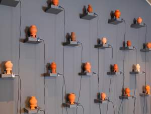 “A Transient's Paradox” is a sculptural installation of 22 rounded clay vases in various tones of terracotta orange and brown with flared legs, each resting on a small gray shelf and spaced out in a staggered grid. On each, a wire extends from the base of the vase, connected to a computer chip on the shelf, and a USB cable extends from that, draping to the floor.