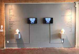 “A Symmetrical Despondency” is a symmetrical sculptural installation centered on two white lidded ceramic vessels on shelves. Connected by cable channels to each of the shelves are tv screens and each screen shows the lower half of a woman’s face in black and white appearing to speak. On the wall behind the left vessel is a poem in English titled “The Story of Vagrancy”. Mirrored on the right side of the other vessel appears the same poem in Farsi.