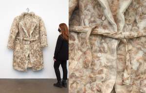 “Sound blanket #10” is a very oversized trenchcoat with white buttons and a belt tied at the waist hanging on a silver hanger on a white wall. The surface is made of swirling felted white and brown wool and hair.