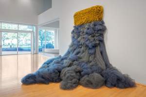 “We Cannot Weep Without Vibrating the Waters” is a sculptural installation on the white wall of a museum space. From a rounded rectangular band of densely ruffled mustard tulle on the wall, a cascading form of bundles of gray and lavender tulle fabric spill down, piling outwards onto the floor below.