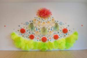 “Lo que la montaña anhela” is a sculptural wall installation, forming a symmetrical upside down fan shape formed of accumulated circles and poms of fabric. The piece radiates from a large gathered tulle pom in oranges-fuschia colors, followed by rows of pom-flowers in yellow, light green, and orange, radiating out from center, ending in a skirting band of gathered neon green-yellow tulle.