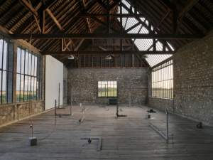 “Untitled” is a sculptural installation in a barn structure with a wooden roof, cobblestone walls, and two large expanses of gridded windows. On the wooden planked floor are staggered concrete sculptures in L forms with irregular edges, framing or holding other objects. Copper tubing extends from several concrete cubes.