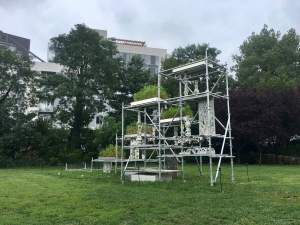“Subterra” is an outdoor sculptural installation formed of a tiered silver scaffolding structure that collects and recycles rainwater. Trays of blooming plants are stacked on the scaffolding, with water draining through irregular columns and planks of pocked concrete.