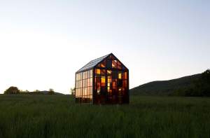 “Solarium” is a greenhouse-like sculpture set on a grassy hill at dusk, formed of gridded panes of yellow, brown, and honey colored glass. Tree saplings are visible through the windows.