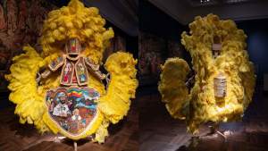 “Ethiopia (Suit)” is a feathered suit standing in a museum display. The central lower apron depicts Haile Selassie, the former Emperor of Ethiopia, warriors, and Ethiopian churches. Arm and chest plates depict other figures, and all medallions are edged with gold velvet ruffles. The voluminous headdress and arms are formed of cascading yellow ostrich feathers.