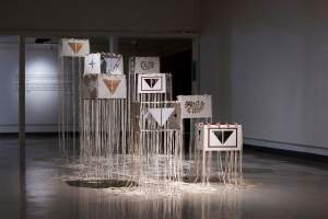 In a darkened gallery room, the sculptural installation “Visitation” stands in the center. Rectangular wide cubes of textured off white handmade paper and clear acrylic float in a staggered group, bound at the edges with tied white deer hide laces - long strings of lacing hang down from the edges and pool on the floor. Various cubes have cut out or etched Lakota motifs of power including paired triangles and 4-point stars.