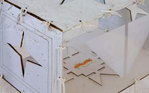 This detail of “Visitation” focuses on a corner of a wide cube that is off white handmade paper on 5 faces, and clear acrylic on the front face. Each paper face has various cut out Lakota motifs, and the clear face has a pair of triangles etched in white. Each face is tied together with white deer hide lacings.