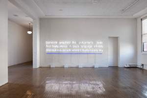 “Landscape V” is an installation of block-justified white neon words in 3 rows, with a neon blue line below the final line, on a wide white wall. The words read “Someone smashed the policeman’s radio And finally silence A black language infinitely And blue in a decade where it finally means sky”.