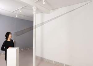 In a white gallery space, a person looks into sculpture “The Portal” which is a very elongated transparent cone made from very finely woven black hair that stretches from their face to the upper corner of the room.