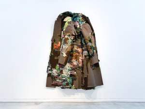 An intangible Sanctuary Of Ocean And Stars II is a sculptural textile work resembling a hanging coat, composed of layered and frayed fabric pieces in earthy and muted tones, displayed against a white wall.