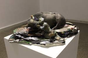 All Her Calves Were Slaughtered is a sculpture depicting a kneeling figure embracing a reclining cow, their faces touching, both constructed from various textured and colored fabrics, displayed on a white pedestal.