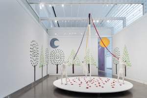 A photo of the Queens Museum's Gallery containing a large installation with three white, life-sized dog sculptures skipping on a white, circular platform and holding lavender, red, and orange ribbons attached to a large white cane pole. The platform is covered in pink, magenta, and red paper mache flowers. The gallery walls are covered with trees made of light and dark green painted paper mache leaves. The gallery also features a grey-blue crescent moon and a large orange sun floating above the back entrance.