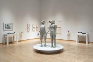 Two sculptures depicting hybrid human-animal figures stand side by side in a gallery space. They hold hands, with their heads slightly turned to gaze affectionately at one another. On the right, is a naked woman with a dog’s head; the figure on the left is an anthropomorphized dog with a long tail standing on its hind legs with the head of a woman.