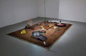 Net Worth is a installation on a soil-covered surface, showcasing assemblages of various objects, including books, a basketball hoop, a framed picture, a model skeleton, and scattered electronic components.