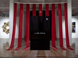 This installation view of Walk with me centers on a large vertical black screen displaying the text "w. Walk with me." in front of a backdrop of strips of red hanging fabric, with other framed artworks visible on the walls.