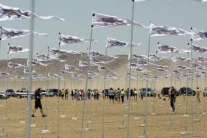 Earth Memory is an environmental art installation with hundreds of triangular banner flags whipping in the wind on silver poles in a dry lakebed in Joshua Tree, CA, with people walking around the installation. Each flag has an image of a pre-historic fish.