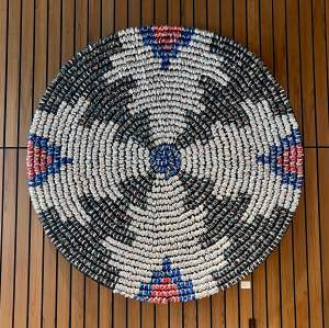 Aerial view of art installation titled Immersion which is a circular medallion with a black symmetrical labyrinth-like design on concentric arcs, with a person in red standing near the edge and sparse vegetation surrounding the area.