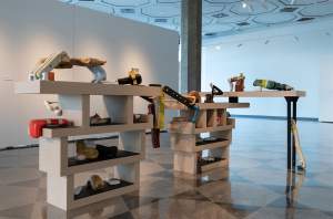 Table of Contents is a sculptural installation in a gallery space featuring various hand tools, biomorphic wooden forms, and objects placed on tiered chunky white shelves