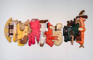Grocery Landscape i﻿s a colorful abstract wall sculpture composed of interconnected, irregularly shaped wooden forms in yellow, pink, red, beige, green, and brown tones, with utilitarian styled accents of metal.