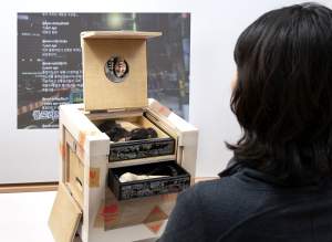 Moonlight in Colorado, Comb box is a sculpture of a small wooden shipping-crate like cube chest, marked with “fragile” stickers. A hinged cover is raised, which holds a small convex mirror, reflecting upside down a viewer standing in front of the piece. Below that, are two shallow drawers with dark mother-of-pearl floral inlay fronts, the top holding a wig of dark and blonde hair, and in the second drawer, a paper microphone. Behind the piece is a projected video showing a feed of social media messages in Korean.