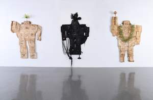 Mamabots - Ms. Daegu, Hindsight 2020, Ma(ma)bot are 3 blocky sculptural figures hanging on a white wall. Each is composed of an assemblage of picture frames, encased in a wrinkly, paper-like skin, with circles in the skin exposing fragments of the pictures inside. The left tan mamabot has a palm perched on its head, and raises one arm. The center mamabot is black, with feathers springing from its head and looped strings hanging from left arm. The right mamabot is tan, has an arm raised with a wand, and a vegetal necklace.