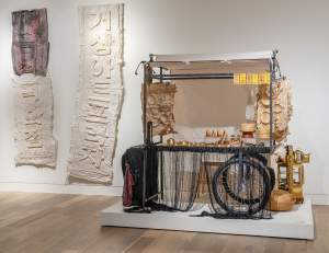 Street Art Cart is a mobile installation reminiscent of a street food cart with two bicycle wheels. On a white plinth, the cart table holds an assortment of brass containers and steamer baskets, paper castings of dishes hang from the back, and a black fringe hangs from the edge. On the wall are two large vertical paper castings of raised Korean letters.