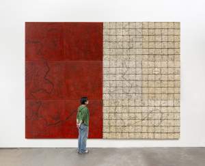 Old Homeplace is a large abstract diptych, more than twice as tall as the person standing in front of it. On left, is a textured red wax panel, with incised dark outlines of rivers and creeks. On the right, is a grid of white maps showing allotment lines in the Cooweescoowee District of Cherokee Nation, railroads, names of people, crudely sewn together with hog rings, overlaid with the same waterways.