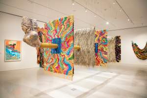 to leave a trace is a sculptural installation of five textile panels suspended from the ceiling, alternately covered with colorful textile weavings or dried grass. A wooden bust of a goddess extends from the front panel, from a trunk that extends through the center of each of the staggered panels.