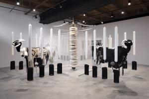 + [pronounced t’] is a sculptural installation of black and white components suspended from the ceiling featuring stylized ram sculptures, rows of tall cylindrical white candle-like forms above black cylinders, extending in iterations from a central vertical structure made of stacked disks.