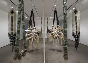 Hólǫ́ llUllUHIbI [duet] is an installation of large hanging sculptures with stacked cylindrical shapes and draped materials, resembling oversized necklaces and beaded forms, displayed symmetrically in a white-walled gallery space.