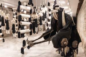 iiZiiT [3]: Riege Jewelry + Supply is a performance and installation in which a person with face obscured in dark regalia and headdress crouches in front of oversized hanging beaded forms made of black and white textiles.