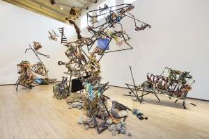 Stream shows 3 abstract sculptures made of metal rods, wood, and mixed materials, forming jumbled and fragmented forms that extend upward in the gallery space.