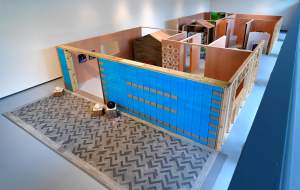 Future Fossils: SUM is an exhibition of a full-scale, single family house cut open and filled with the “sum” of things consumed and disposed of in a human lifetime. Viewed from above, with no ceiling, walls are framed in wood and blue and grey plastic containers, and artworks hang on the wall.