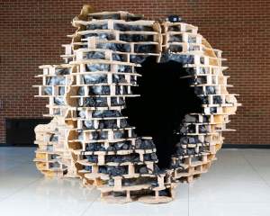 A roughly ball-shaped, free-standing sculpture is made up of a framework of wood planks and short beams like a blocky honeycomb. The spaces are filled with black asphalt or fabric that creates a sheen like dark mother-of-pearl.
